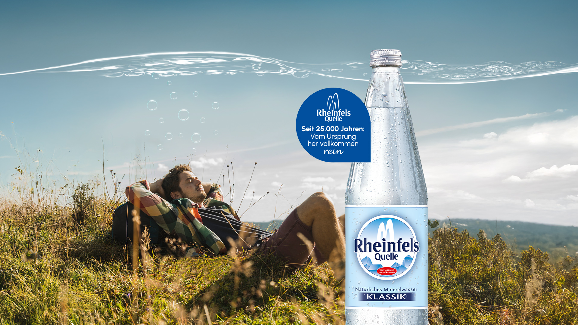 Wanderer liegt im Gras und ruht sich aus. Daneben eine Flasche Rheinfels Quelle Klassik