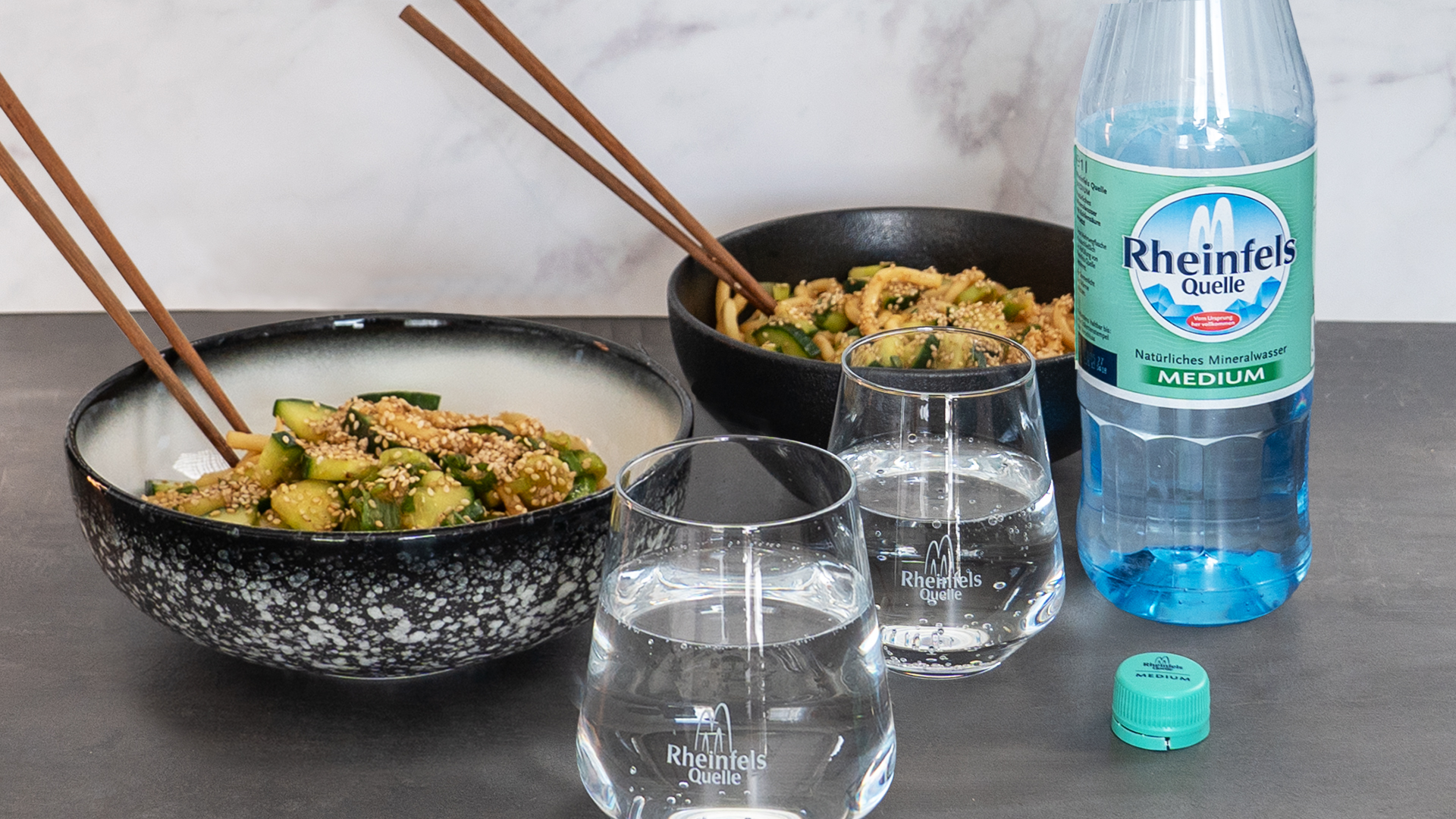 Spicy Udon mit Gurke und Sesam mit einer Flasche Rheinfels Quelle Medium auf einem Holzbrett in Schüssel serviert