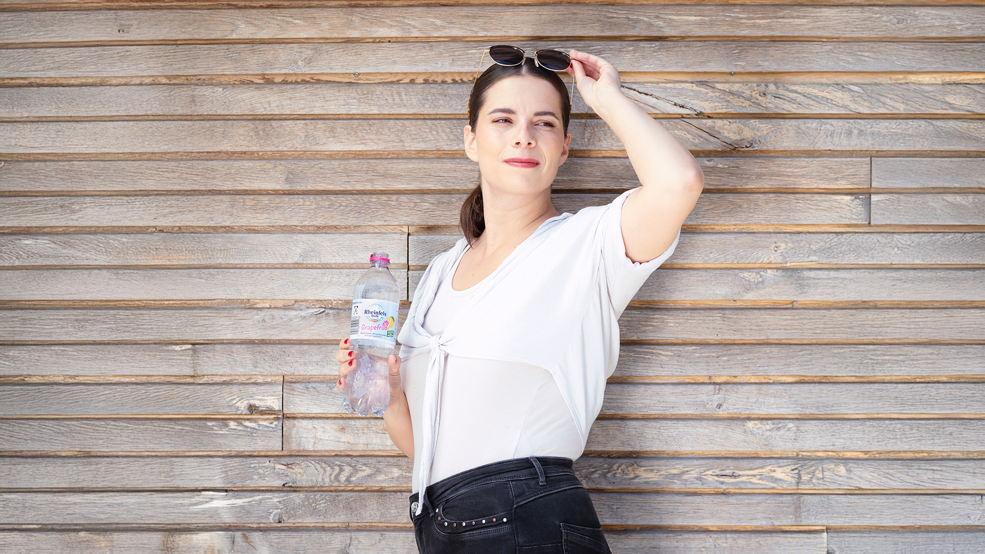 Eine junge Frau lehnt an einer Holzwand und hat eine geöffnete Flasche Rheinfels Quelle Grapefruit in der Hand.