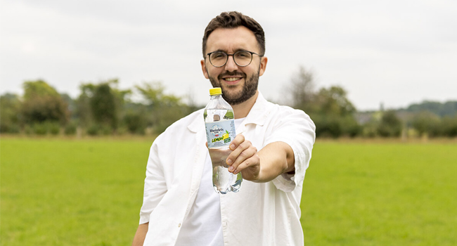 Ein Mann steht auf einer Wiese und hält lächelnd eine Flasche Lemon in die Kamera.