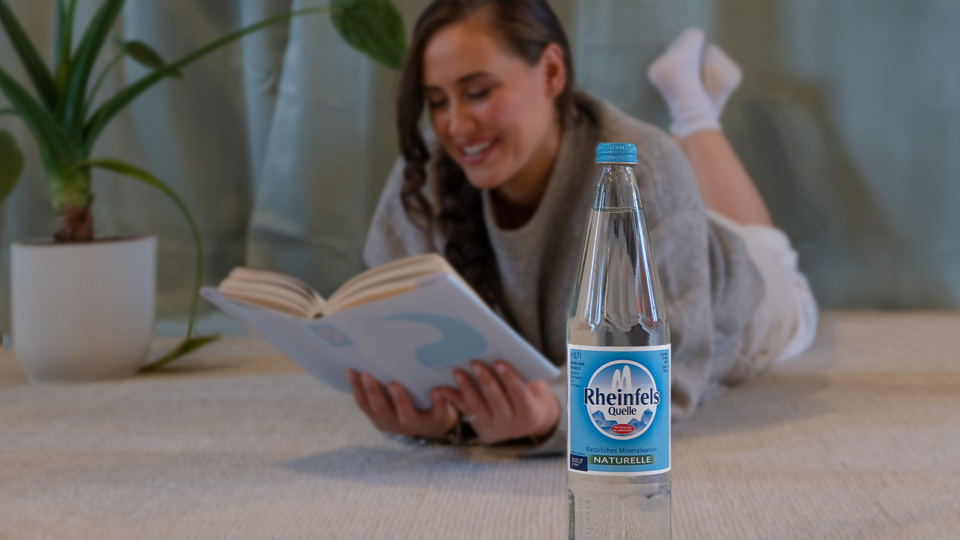 Eine Frau liegt auf einem Teppich und liest, eine Glasflasche Rheinfels Quelle Naturelle steht neben ihr.